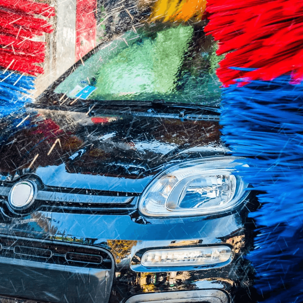 Auto in der Waschanlage, umgeben von rot-blau-gelben Bürsten und Wasserstrahlen während der Reinigung.