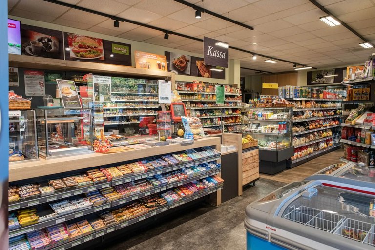 Breitwinkelaufnahme des Nah & Frisch Shops in der Tankstelle Loibl, mit gut sortierten Regalen voller Süßwaren, Snacks, Getränken und Tiefkühlprodukten; im Vordergrund die Kassa mit Frischwarenbereich und Kühltruhe.