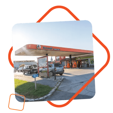 Tankstelle der Marke Turmöl bei Sonnenschein, mit mehreren Autos an den Zapfsäulen und einem gut sichtbaren Shop im Hintergrund; das Bild ist leicht schräg eingerahmt von einem orangen Grafikelement.
